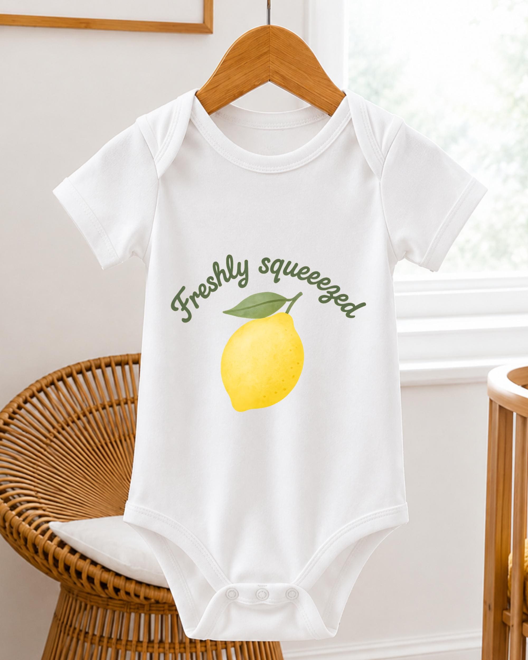 A white baby onesie with a lemon graphic and the text "Freshly squeezed" printed on it, hanging on a wooden hanger.
