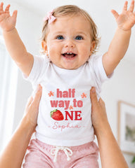 A smiling baby girl wearing a white t-shirt with the text "half way to one" and a strawberry graphic, being held up by someone's arms.