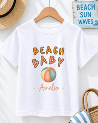 A white t-shirt with the text "BEACH BABY" printed on it, along with an illustration of a beach ball. The t-shirt is hanging on a wooden hanger, and there is a straw hat and a blue sign with the text "BEACH SUN WAVE" displayed in the background.