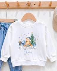 A white sweatshirt with a Christmas-themed design, including a teddy bear, presents, and a tree, along with the text "My first Christmas Oscar".