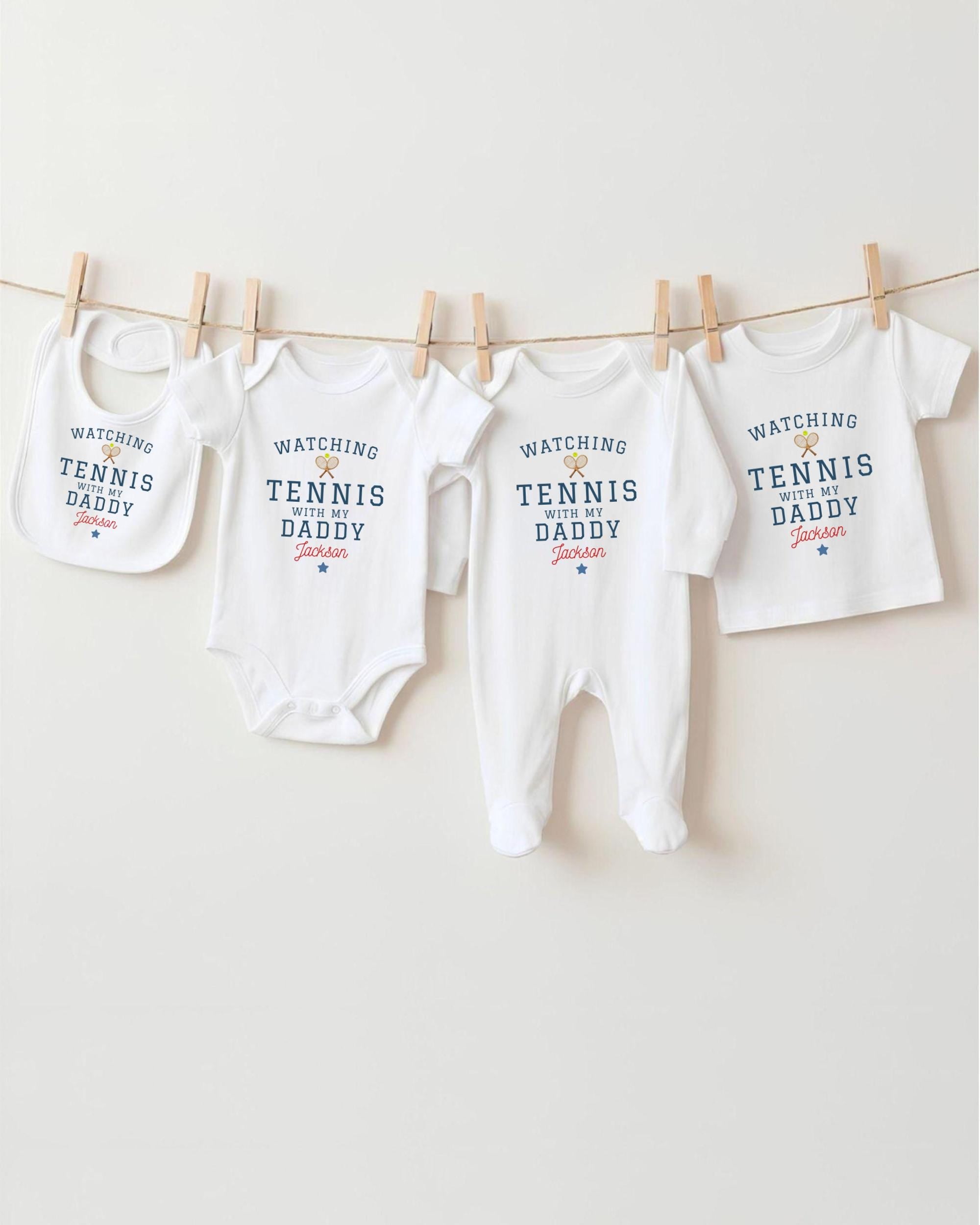 A row of four white baby clothes with the text "WATCHING TENNIS DADDY Jackson" printed on them, hanging on a clothesline.