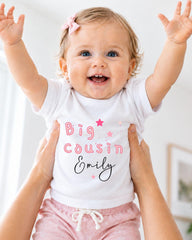 A smiling baby girl wearing a white t-shirt with the text "Big Cousin Emily" and pink stars, being held up by someone's arms.