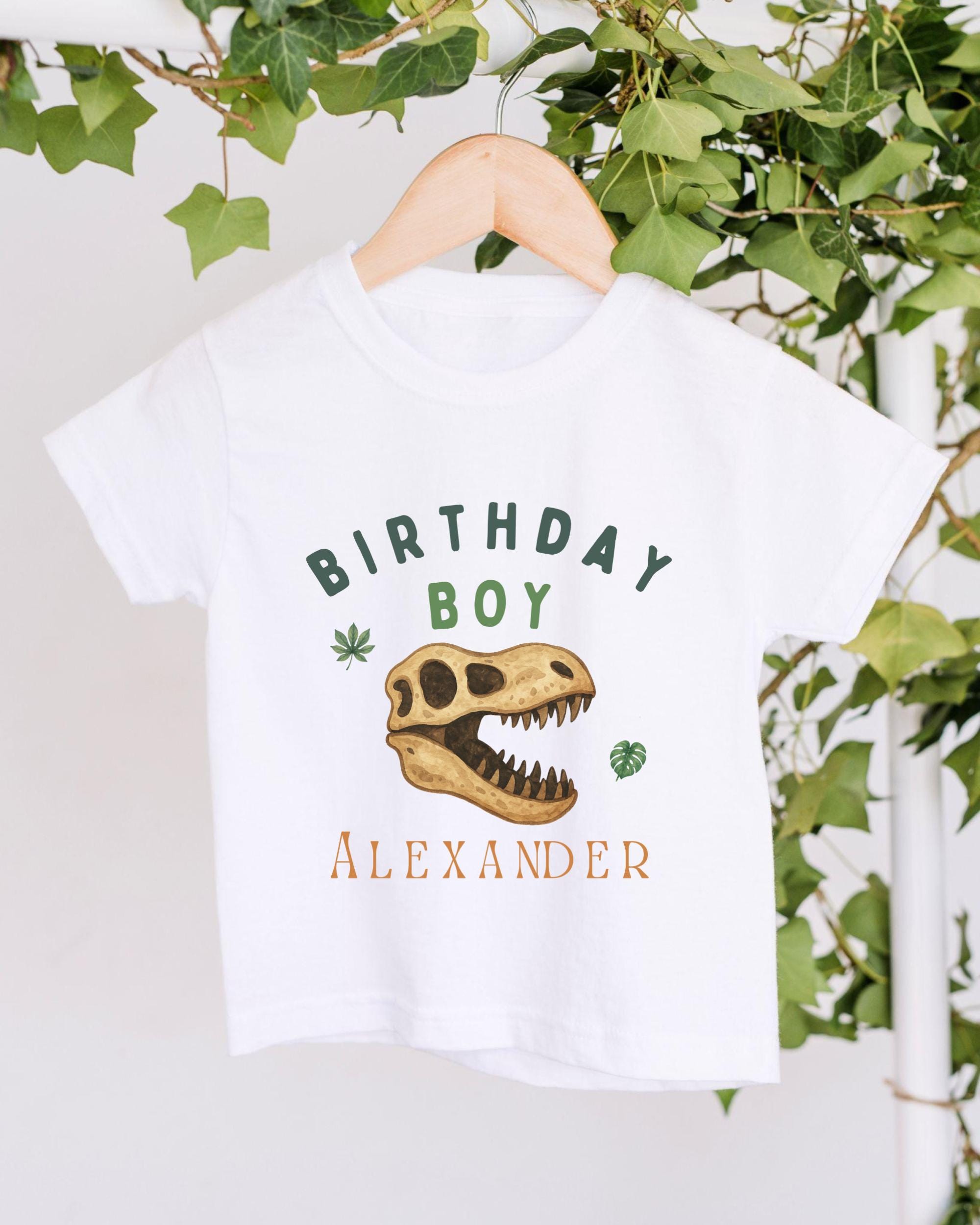 A white t-shirt with a dinosaur skull and the text "BIRTHDAY BOY ALEXANDER" printed on it, hanging on a wooden hanger.