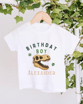 A white t-shirt with a dinosaur skull and the text "BIRTHDAY BOY ALEXANDER" printed on it, hanging on a wooden hanger.
