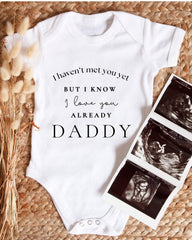 A white baby onesie with text printed on it, placed next to a set of black and white ultrasound images.