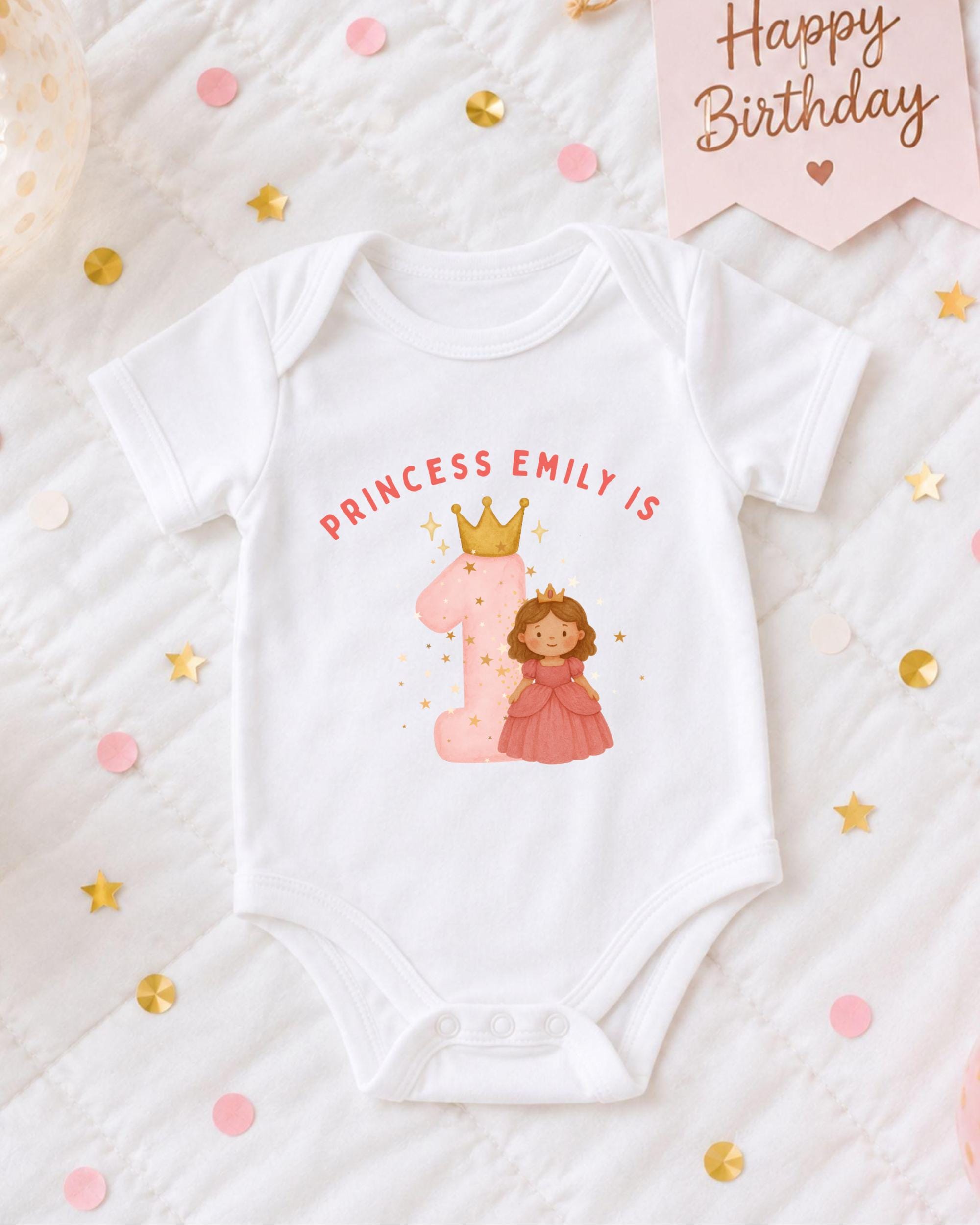 A white onesie with a princess and a crown, along with the text "PRINCESS EMILY IS" and a number 1, is displayed on a bed with confetti and a birthday card.
