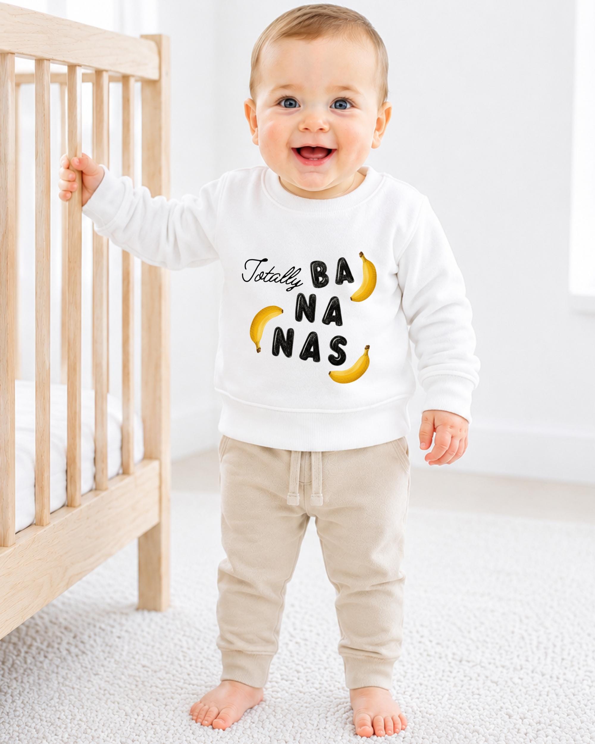 A smiling baby boy wearing a white sweatshirt with the text "Totally BANANA NAS" printed on it, standing next to a wooden crib.