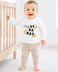 A smiling baby boy wearing a white sweatshirt with the text "Totally BANANA NAS" printed on it, standing next to a wooden crib.