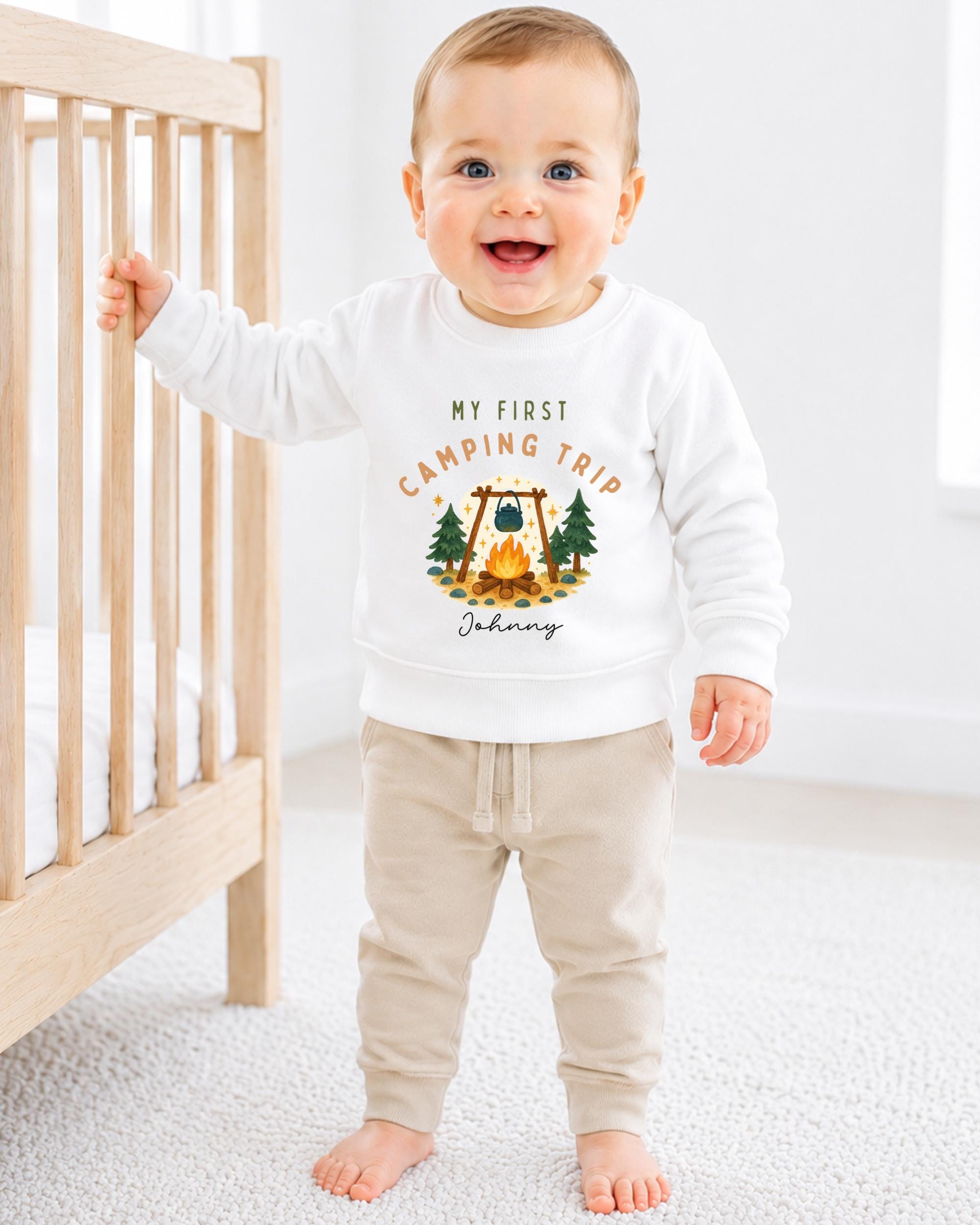 A smiling baby boy wearing a white sweatshirt with a camping trip design and the text "My first camping trip" is standing next to a wooden crib.