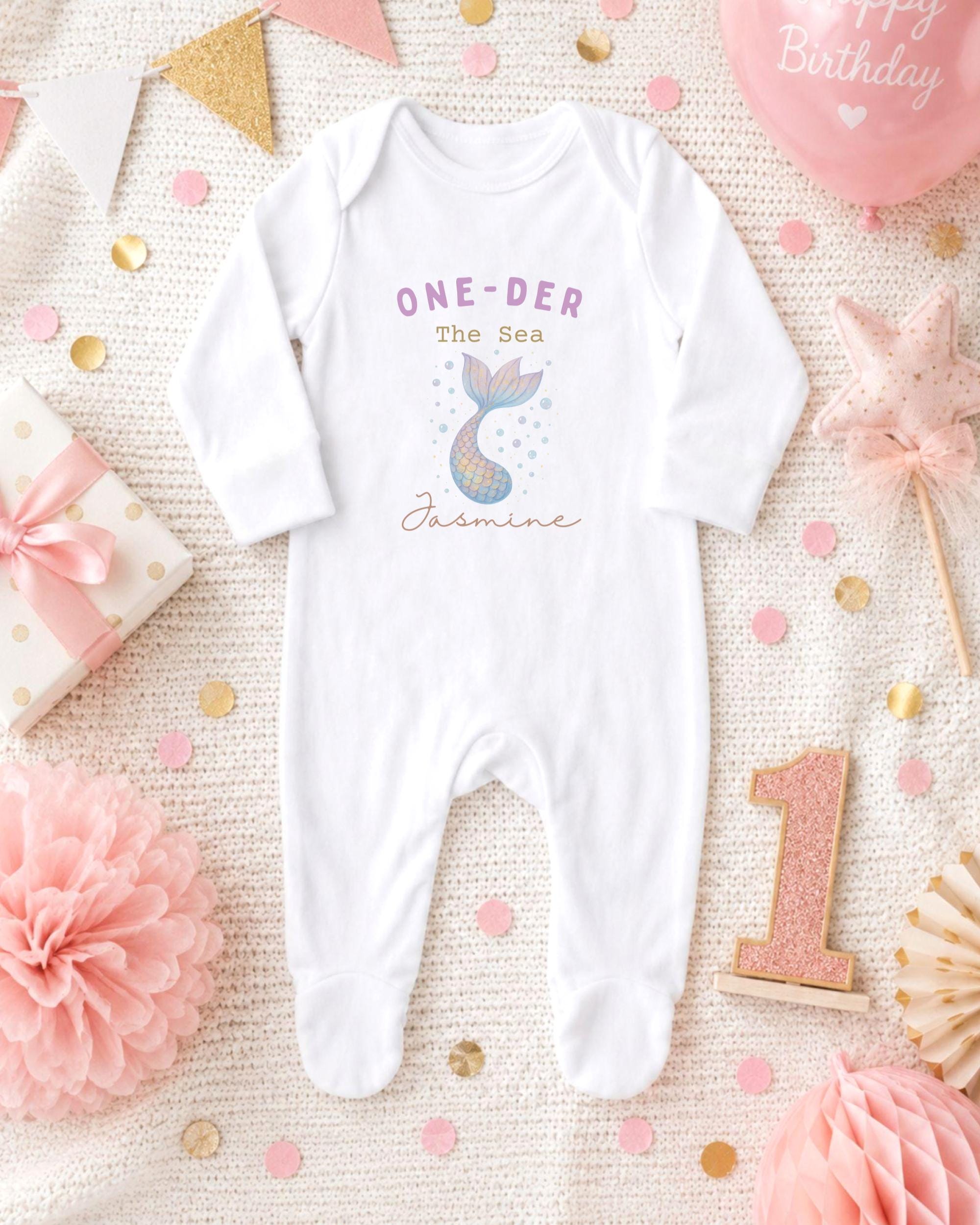A white onesie with a mermaid design and the text "One-DER The Sea Jasmine" printed on it, surrounded by pink and gold confetti, a pink star, and a pink ribbon.