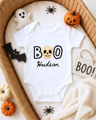 A white onesie with the text "BOO Hudson" printed on it, surrounded by Halloween-themed decorations such as a ghost, a pumpkin, a bat, and a sign that says "BOO!".