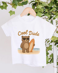 A white t-shirt with a cartoon image of a bear holding a surfboard and wearing sunglasses, with the text "Cool Dude" printed on it.