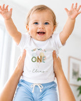 A smiling baby wearing a white t-shirt with the text "Half Way to One - Elias" is being held up by someone's arms.