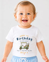 A smiling baby boy wearing a white t-shirt with a golf cart graphic and text that reads "Happy 1st Birthday as my Daddy You're The Best By Par Ollie x".