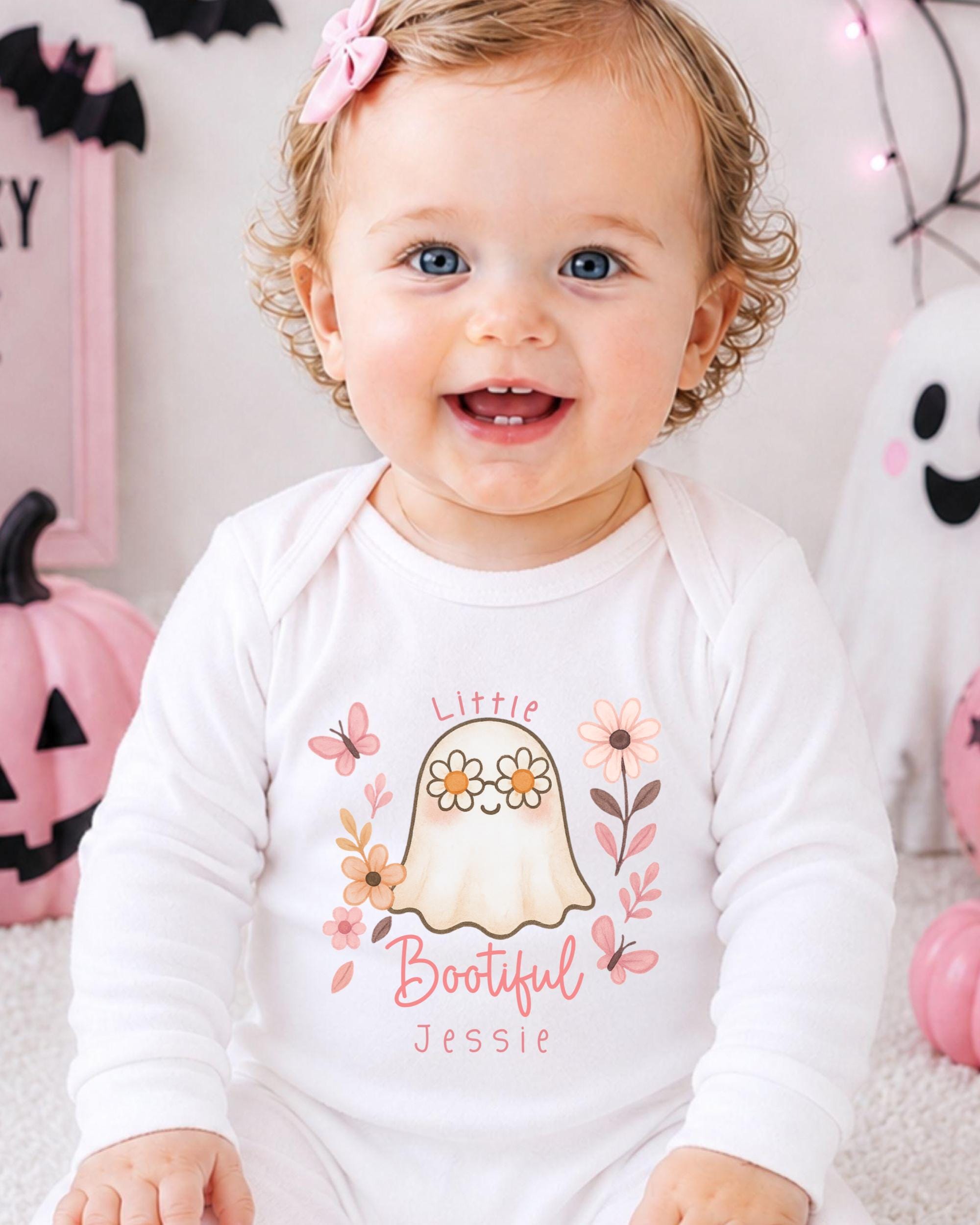 A smiling baby girl wearing a white onesie with a ghost and floral design, sitting in front of Halloween decorations.