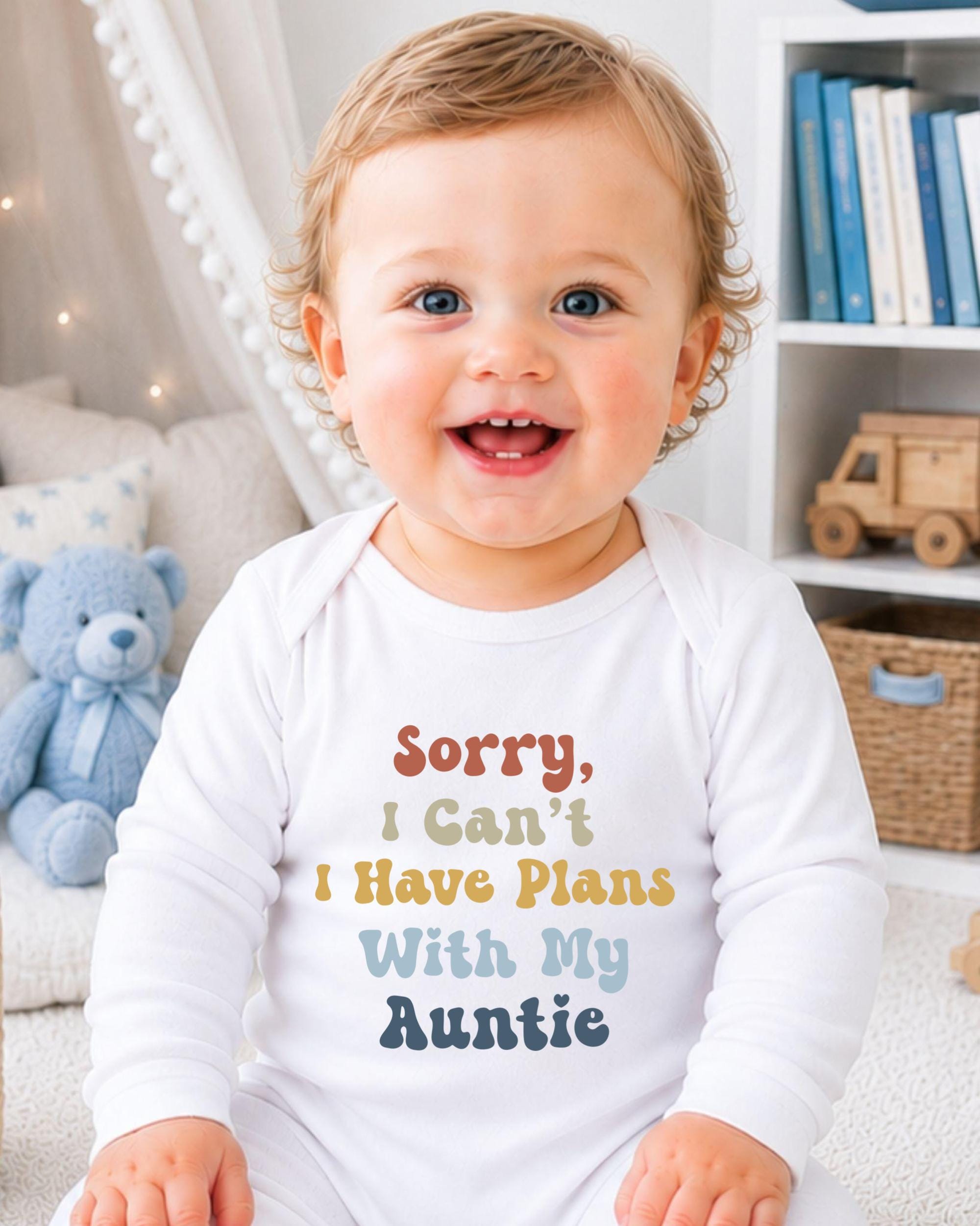A smiling baby wearing a white onesie with text that says "Sorry, I can't I have plans with my auntie" is sitting in a room with a teddy bear and bookshelf in the background.