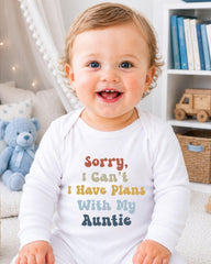 A smiling baby wearing a white onesie with text that says "Sorry, I can't I have plans with my auntie" is sitting in a room with a teddy bear and bookshelf in the background.