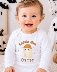 A smiling baby boy wearing a white onesie with a cartoon ghost and the text "Little Boo Oscar" is sitting in front of Halloween decorations.