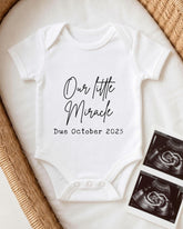 A white baby onesie with the text "Our little Miracle Due October 2015" printed on it, placed on a bed with a woven basket. There are also two black and white images of an Ultrasound scan, which appear to be related to the announcement of the baby's arrival.