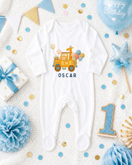A white onesie with a giraffe and balloons, surrounded by birthday decorations such as a blue balloon, blue and gold confetti, and a blue ribbon.