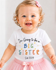 A smiling baby girl wearing a white t-shirt with text and a pink bow.