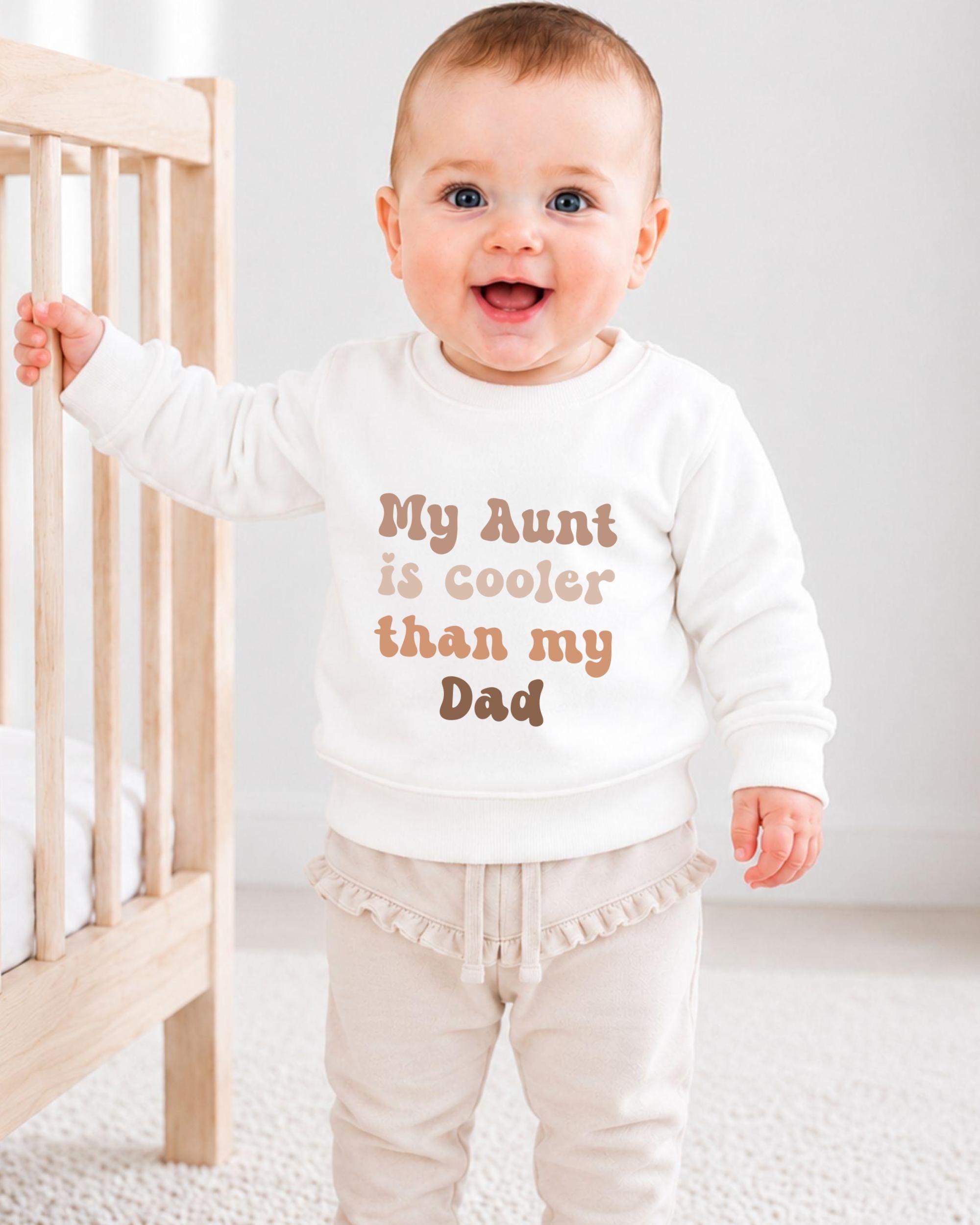 A smiling baby wearing a white sweatshirt with the text "My Aunt is cooler than my Dad" printed on it.