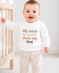 A smiling baby wearing a white sweatshirt with the text "My Aunt is cooler than my Dad" printed on it.
