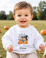 A smiling baby boy wearing a white sweater with a teddy bear and pumpkins, sitting in a field with pumpkins in the background.
