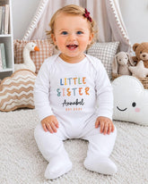 A smiling baby girl wearing a white onesie with the text "Little Sister Annabel" sits on a white carpet surrounded by stuffed animals and pillows.