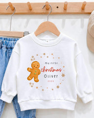 A white sweater with a gingerbread man graphic and text that says "My first christmas Oliver" printed on it.