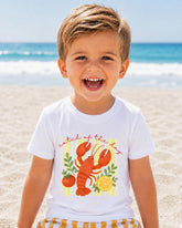 A young boy with a big smile on his face, wearing a white t-shirt with a lobster graphic and the text "Catch of the day" printed on it, standing on a sandy beach with the ocean in the background.