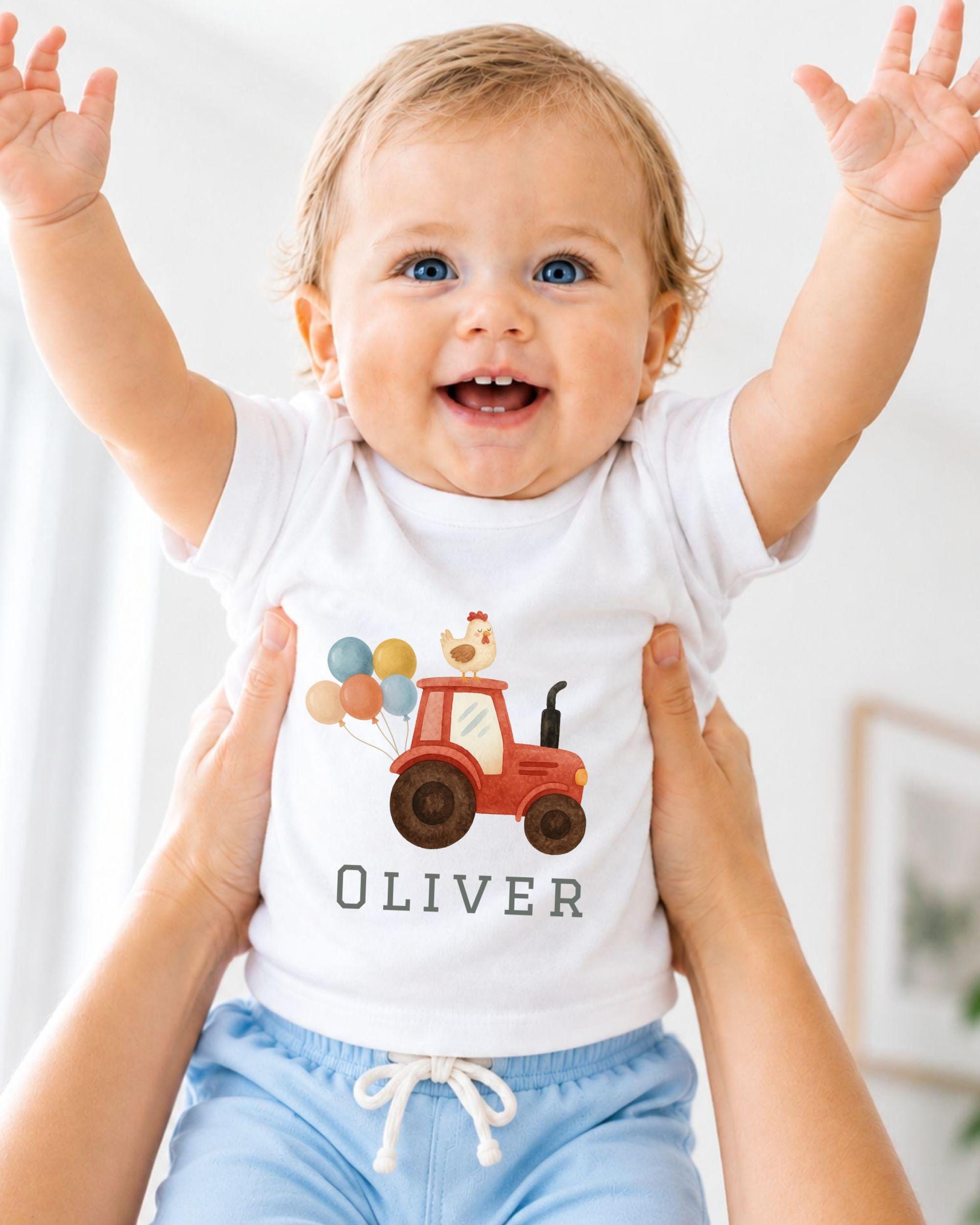 A smiling baby wearing a white t-shirt with a tractor and a chicken on it, being held up by someone's arms.