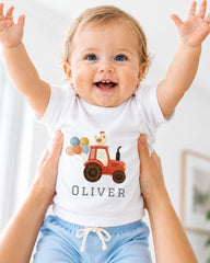 A smiling baby wearing a white t-shirt with a tractor and a chicken on it, being held up by someone's arms.