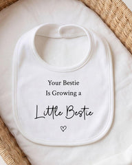 A white bib with the text "Your Bestie Is Growing a Little Bestie" printed on it, placed on a white cloth.