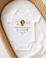 A white baby onesie with a personalized message and a crown on it, placed on a white blanket.