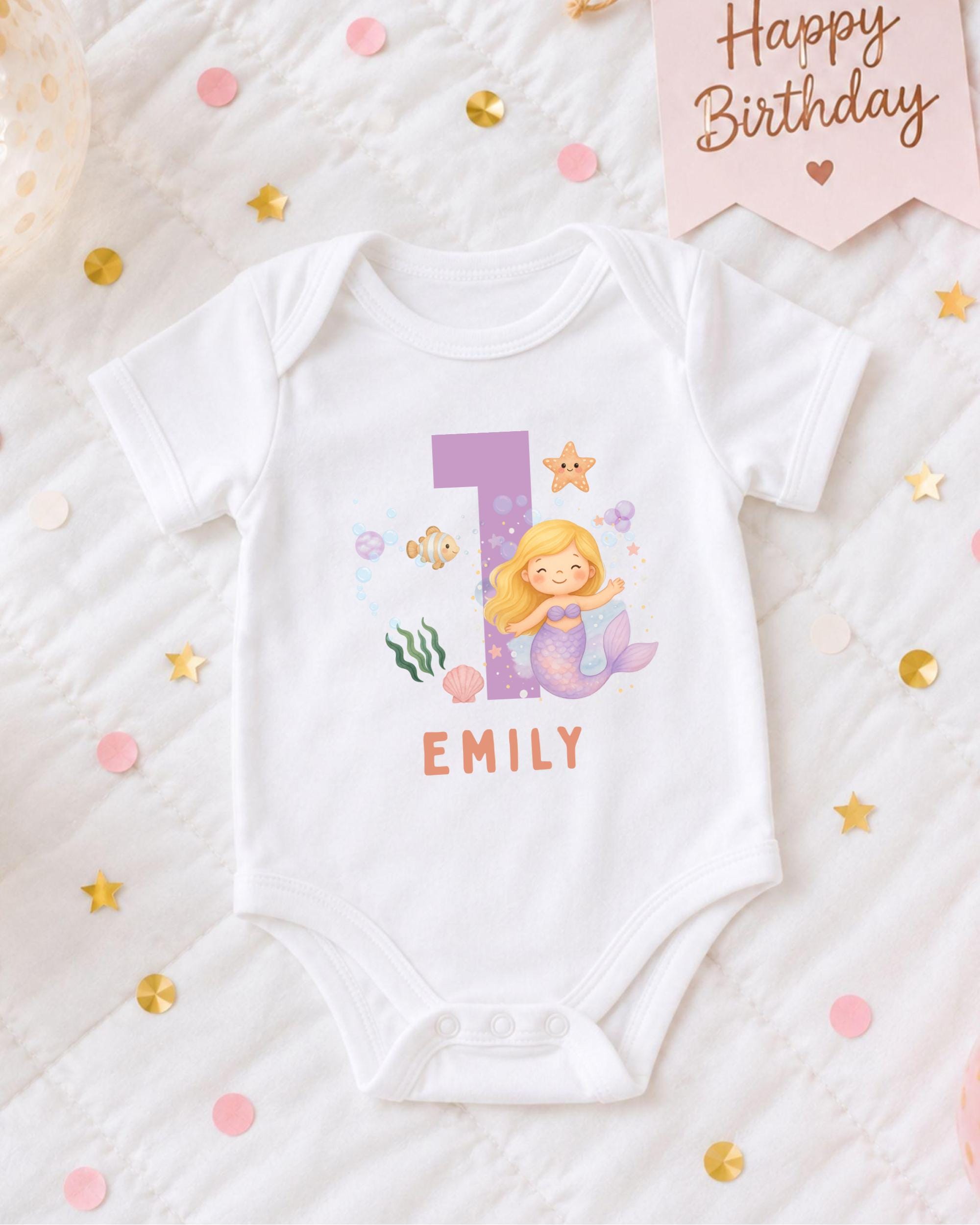 A white onesie with a mermaid design and the number 1, along with the name "EMILY" printed on it, is displayed on a bed with confetti and a birthday card.