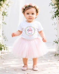 A young girl wearing a white t-shirt with a floral design and a pink tulle skirt, standing in front of a white arch with flowers.