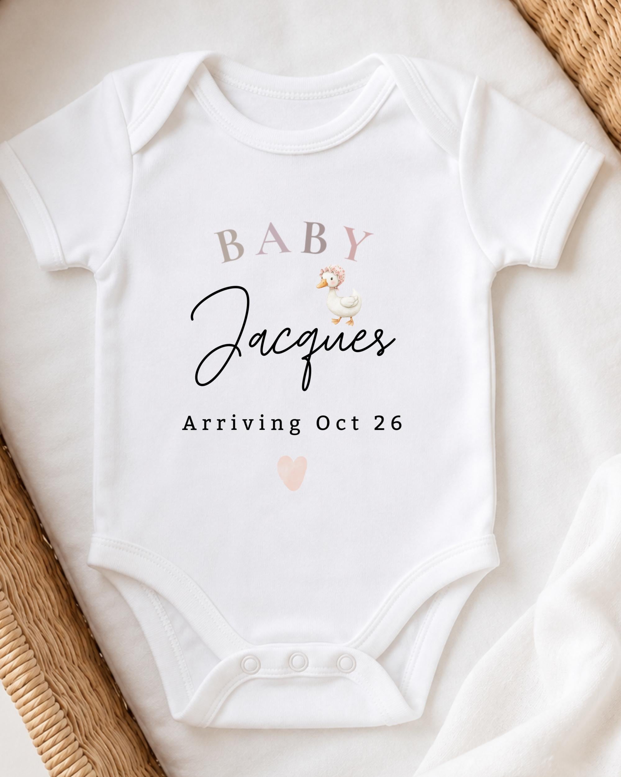 A white baby onesie with text and a small cartoon duck printed on it, placed on a white surface.