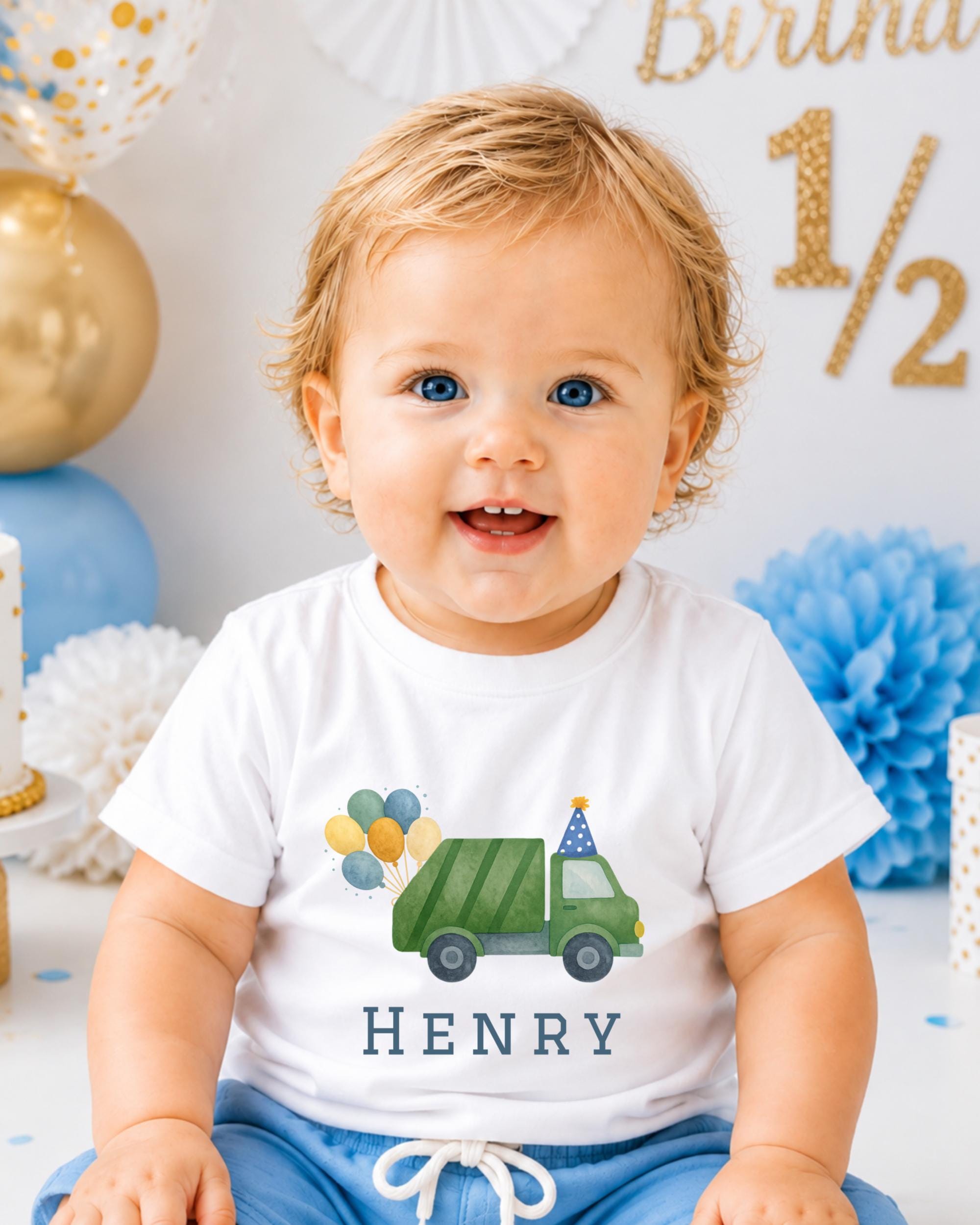 A smiling baby boy wearing a white t-shirt with a green truck graphic and the name "HENRY" printed on it, sitting in front of a birthday party backdrop.