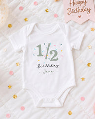 A white onesie with the text "1/2 Birthday - Jane" printed on it, along with a pink banner that says "Happy Birthday" and confetti scattered around.