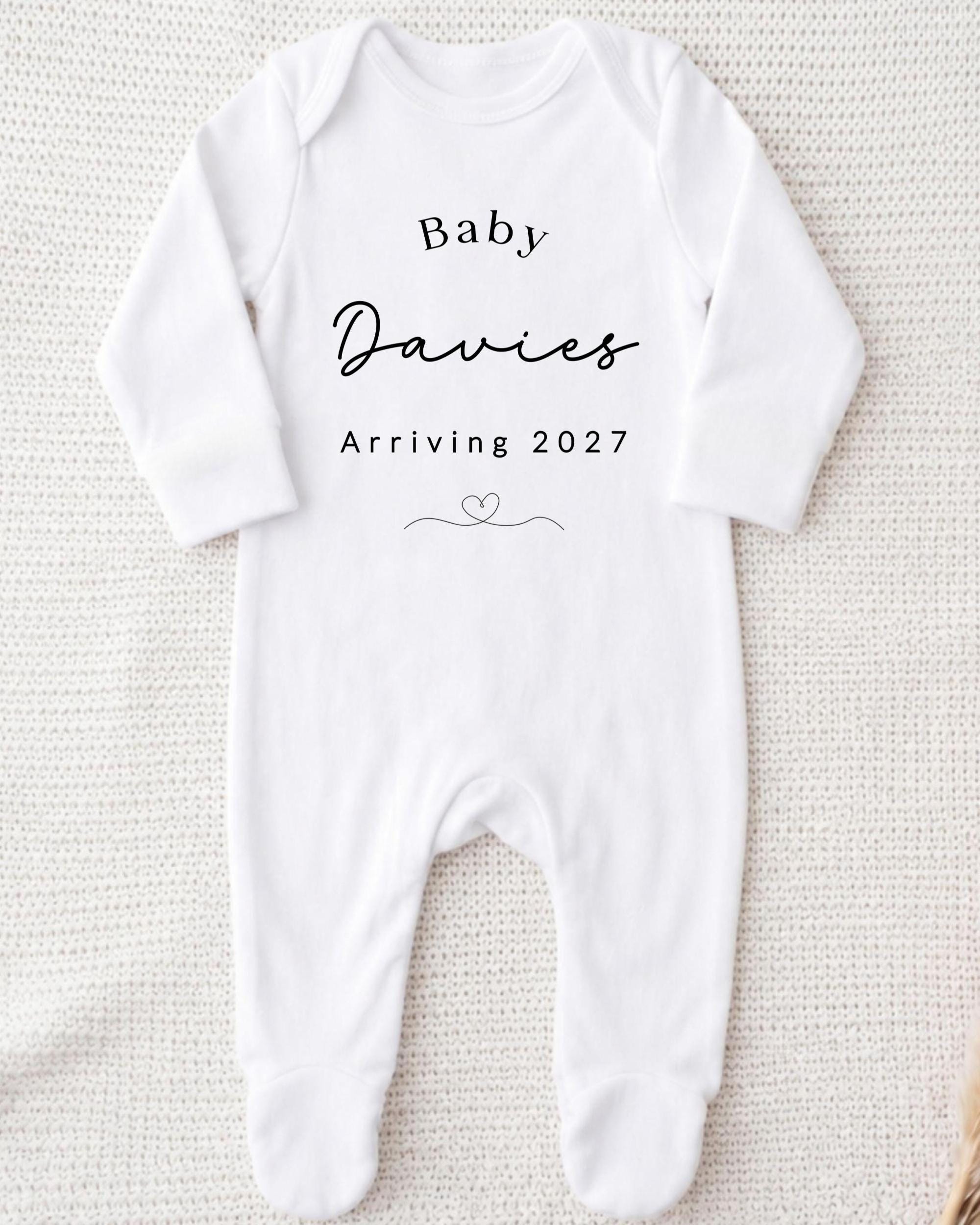 A white onesie with the text "Baby Davies Arriving 2027" printed on it.