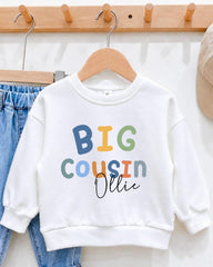A white sweatshirt with the text "BIG COUSIN OLLIE" printed on it, hanging on a wooden hanger.