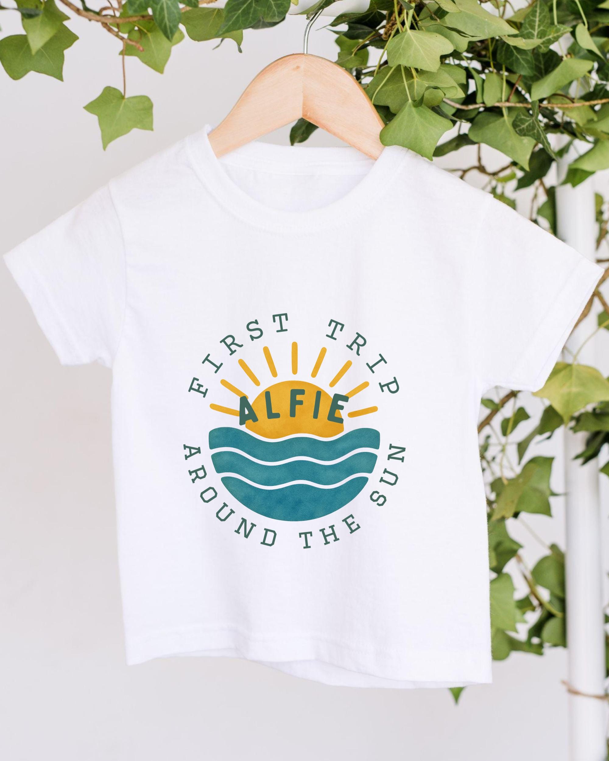 A white t-shirt with a graphic design featuring a sun and waves, along with the text "First Trip Around the Sun" printed on it.