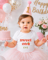 A young girl wearing a birthday hat and a white t-shirt with the text "Sweet on Eve" is posing in front of a birthday cake and balloons.