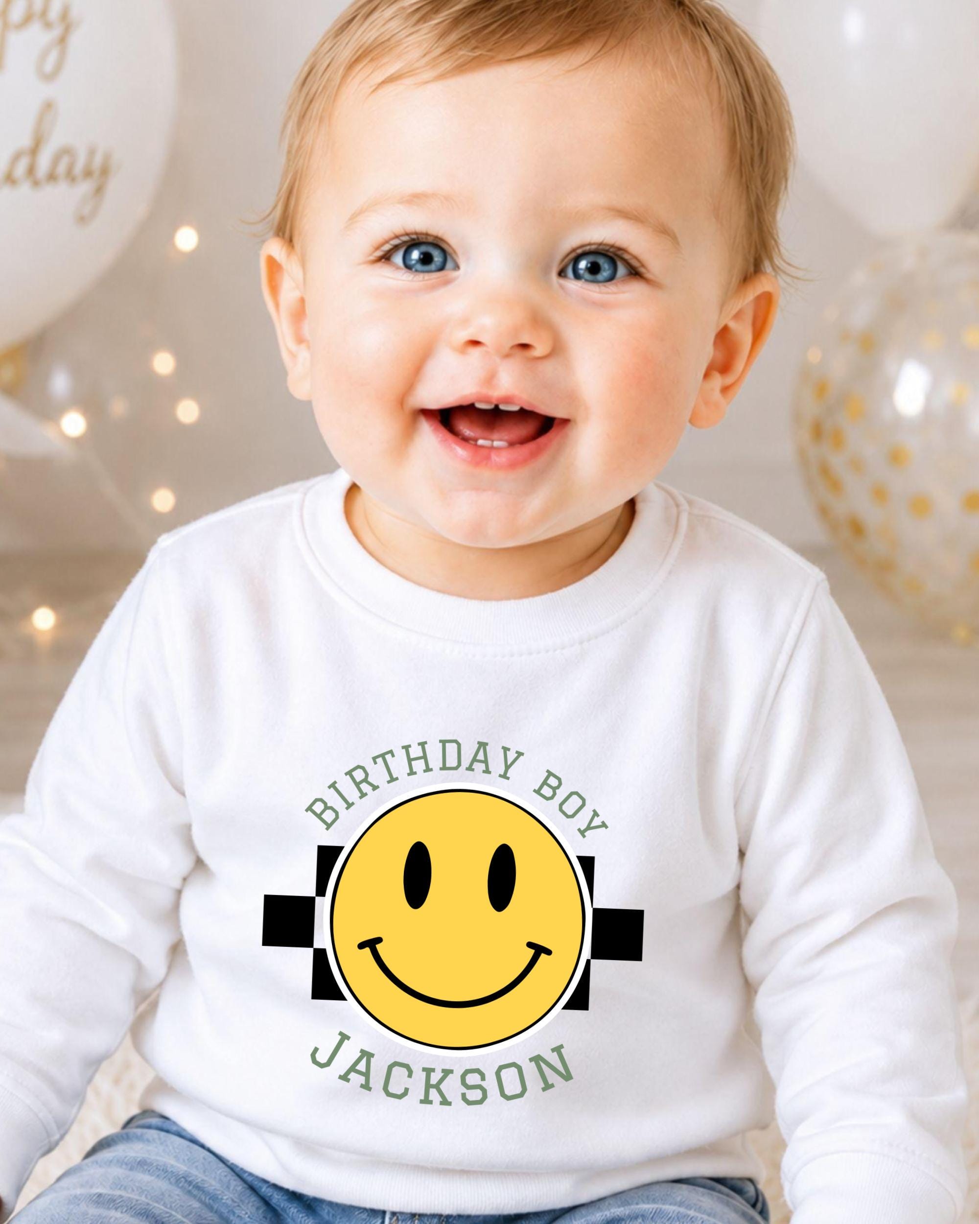 A smiling baby boy wearing a white shirt with a yellow smiley face and text that says "Birthday Boy Jackson".