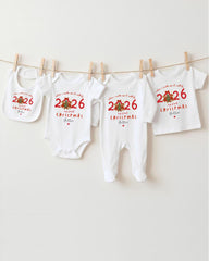 A row of white Christmas-themed baby clothes, including onesies and bibs, hanging on a clothesline.