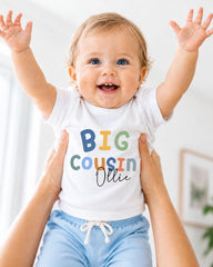 A smiling baby wearing a white t-shirt with the text "BIG COUSIN OLLIE" is being held up by someone's arms.