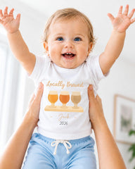 A smiling baby wearing a white shirt with a graphic of three beer glasses and the text "Locally Brewed Jack 2026" is being held up by someone's arms.