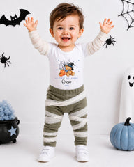 A smiling baby boy wearing a Halloween-themed outfit with a spider and pumpkin decoration, standing in front of a Halloween-themed background.