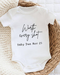 A white baby onesie with the text " Worth every shot Baby Due Nov 28" printed on it, placed on a woven basket.